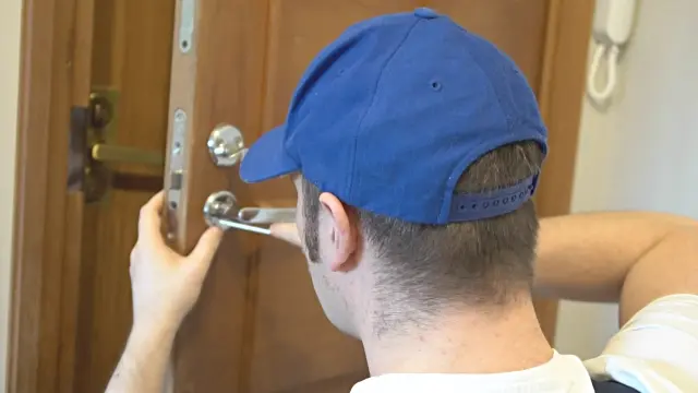 Young handyman in uniform fixing door lock.